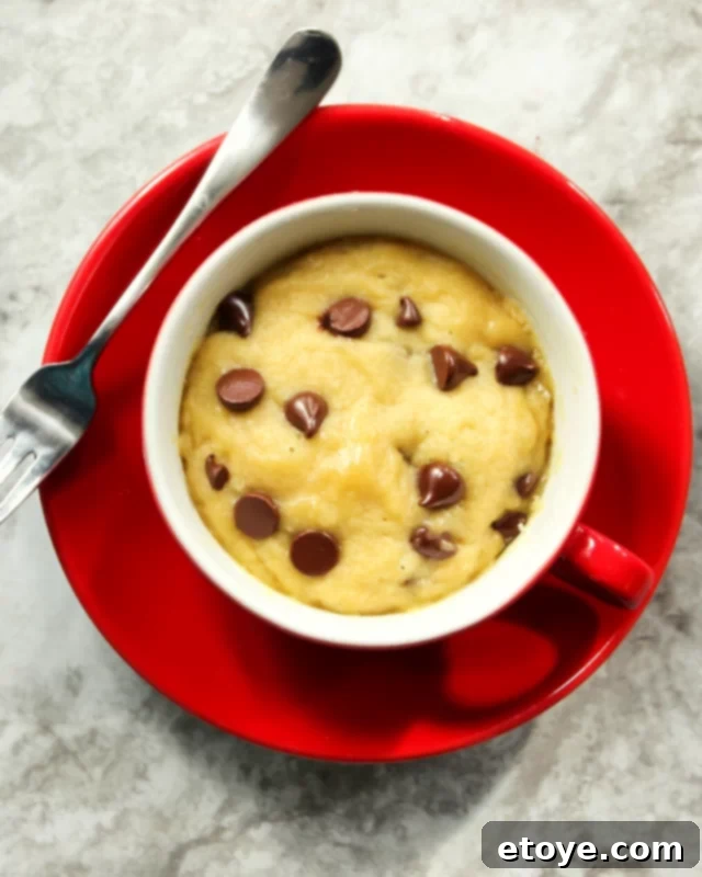 Delicious single-serve chocolate chip cookie in a red mug, placed on a red plate with a fork beside it.