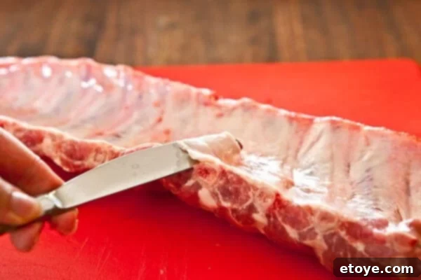 Fall-Off-The-Bone Sweet Chili Ribs 5 A butter knife being used to start lifting the membrane from the baby back ribs, demonstrating the initial step of removal.