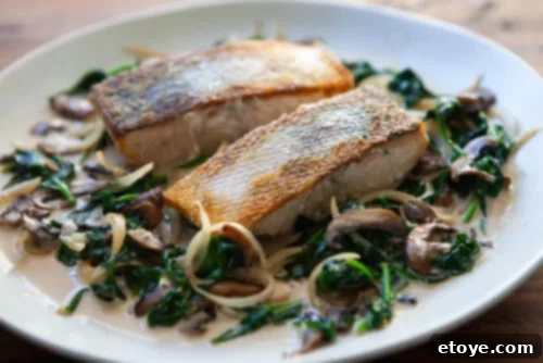 A large skillet filled with creamy mushroom and spinach sauce, with seared salmon fillets nestled within, during the final cooking stage.