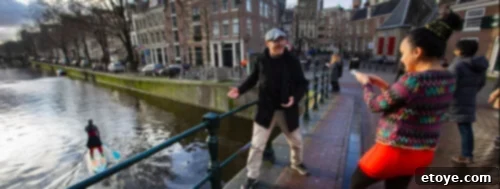 A picturesque canal in Amsterdam with historic buildings lining the sides. A person paddleboards on the water, while on a bridge, a tour guide animatedly talks to a woman wearing a vibrant patterned sweater, capturing the essence of local virtual tours.