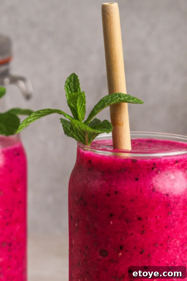 Dragon Fruit Hibiscus Coconut Refresher 3 Close-up of a Dragon Fruit Chill Smoothie in a sleek glass with a reusable straw, showcasing the vibrant pink color and smooth texture, set against a blurred tropical background.