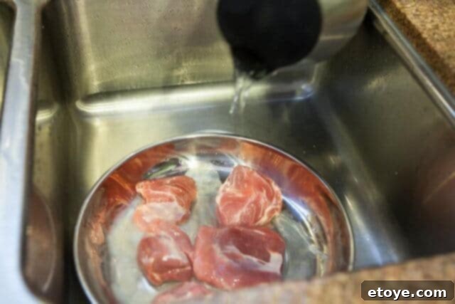 Boiling water being poured over raw pork in a shallow dish as part of a quick par-boiling method.