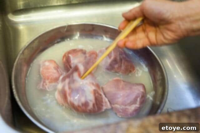 A hand stirring pork pieces in a bowl with hot water, helping to loosen impurities for a clear broth.