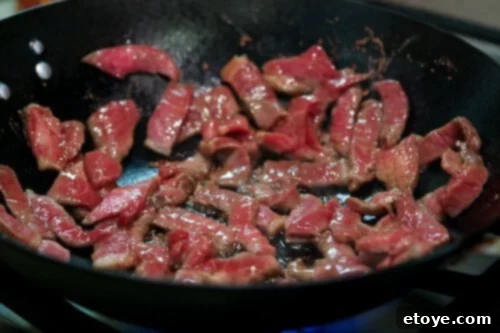 Thinly sliced marinated beef searing in a single layer in a hot wok, showing early signs of caramelization