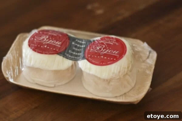 Close-up of a goat cheese round with a purple Thai basil flower bud on top, next to a toasted baguette slice.